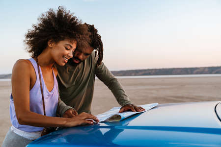 Image of joyful african american couple examining map while travelling with car on desert