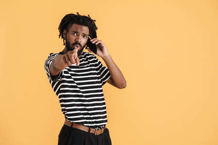 Photo of confused african american guy talking on cellphone and pointing finger at camera isolated...