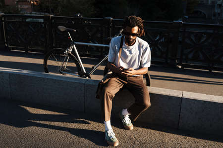 Concentrated african american guy using cellphone while sitting at city streetの写真素材