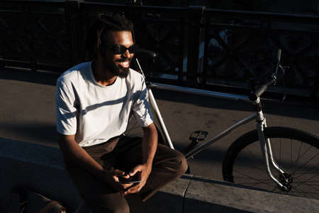 Happy african american guy smiling and using cellphone while sitting at city streetの写真素材