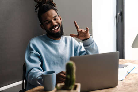 Happy young african man freelancer on a video call while sitting with laptop computer at the deskの写真素材