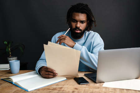 Handsome serious young african man sitting at a coffee shop with pensive look, thinking of business plans, working on laptopの写真素材