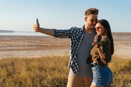 Couple gesturing peace sign and taking selfie on cellphone while strolling on nature at summer