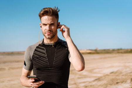 Portrait of a fit sportsman listening to music with earphones while standing at the beach, holding mobile phoneの写真素材