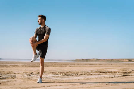 Motivated focused young sportsman exercising at the beach, stetching