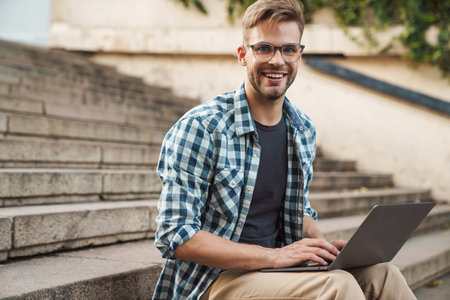 Young man with a laptop on the street, sitting on the stairs working online and communicating with friends via video callingの写真素材
