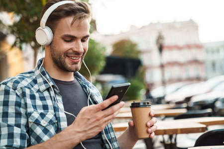 Smiling young man wearing headphones sitting at the cafe table outdoors, using mobile phoneの写真素材