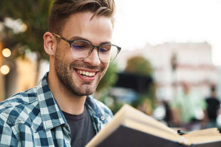 Smiling man enjoying free time and reading a book while sitting outdoors at coffee shop
