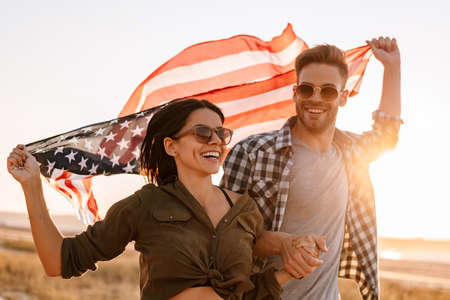 Nice happy couple holding hands together while strolling with american flag on nature at summer