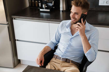 Image of happy positive young businessman sitting indoors at home at the kitchen and talking by...