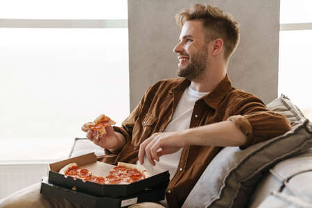Handsome cheerful guy eating pizza while sitting on couch at home