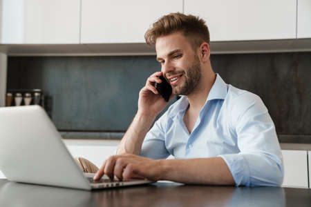 Image of positive young businessman sitting indoors at home at the kitchen and talking by mobile phone while using laptop computerの写真素材