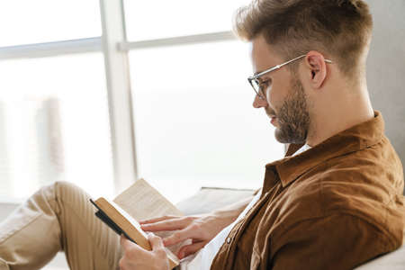 Handsome focused guy in eyeglasses reading book while sitting on sofa at homeの写真素材