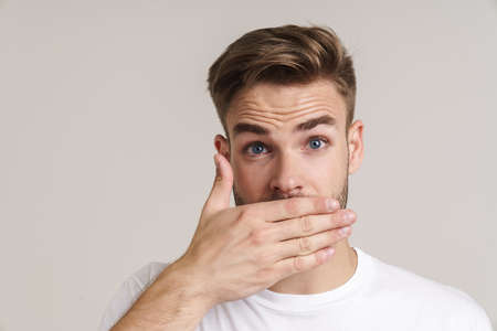 Surprised handsome unshaven guy covering his mouth isolated over grey background