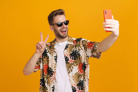 Cheerful handsome guy gesturing peace sign while taking selfie on cellphone isolated over yellow...