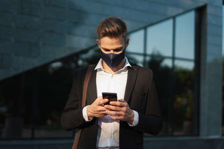 Handsome blonde businessman in face mask using mobile phone on city streetの写真素材