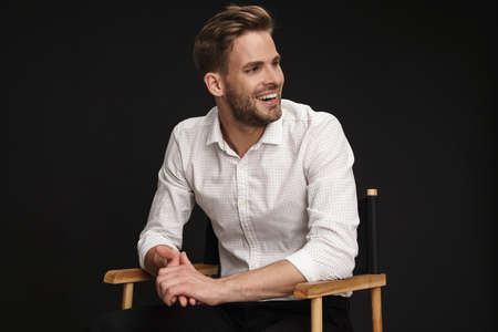 Handsome joyful businessman laughing while sitting in armchair isolated over black background