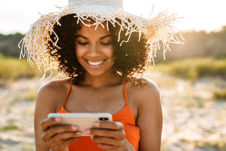 Image of pretty cheery young african woman watching videos by mobile phone at the beach outsideの写真素材