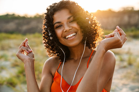 Image of amazing optimistic young african woman listening music with headphones at the beach outsideの写真素材