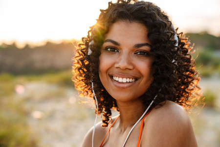 Image of beautiful young african pleased woman listening music with headphones at the beach outside