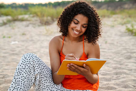Image of pretty optimistic young african woman writing notes in notebook at the beach outsideの写真素材