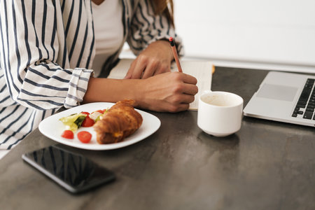 Young beautiful caucasian woman writing in notebook while having breakfast at homeの写真素材