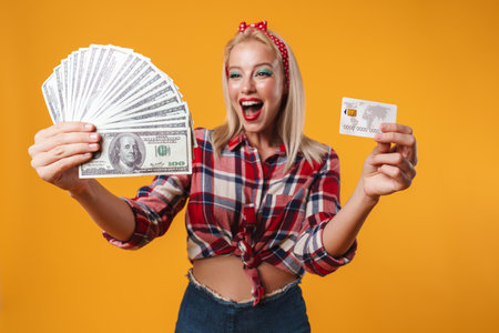 Image of excited blonde pinup girl showing dollars and credit card isolated over yellow backgroundの写真素材