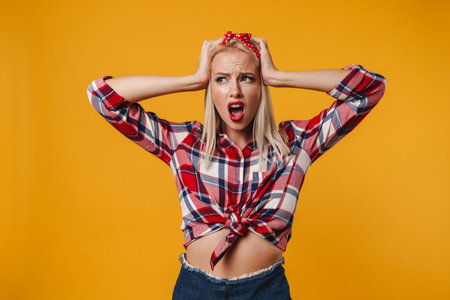 Image of unhappy caucasian pinup girl screaming while grabbing her head isolated over yellow backgroundの写真素材