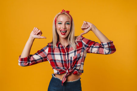 Image of cheerful beautiful pinup girl smiling and pointing fingers at herself isolated over yellow backgroundの写真素材