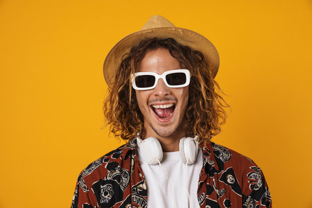Image of cheerful young man in sunglasses and hat laughing at camera isolated over yellow backgroundの写真素材