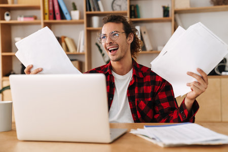 Handsome smiling guy in eyeglasses working with laptop and papers while sitting at officeの写真素材
