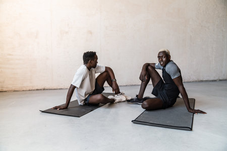 Two young handsome african american men sitting on exercise mats while doing workout indoors