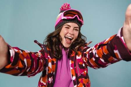 Cheerful young woman wearing snowboard gear taking a selfie isolated over blue backgorund