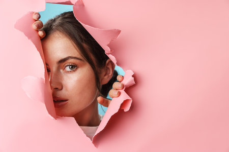 Beautiful young attractive girl peeping through hole in paper isolated over backgroundの写真素材