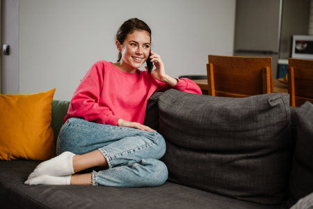 Beautiful joyful girl talking on cellphone and smiling while sitting on couch at homeの写真素材