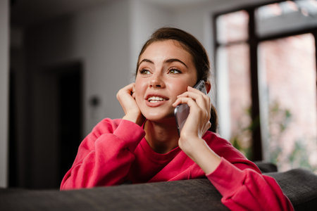 Beautiful joyful girl talking on cellphone and smiling while sitting on couch at home