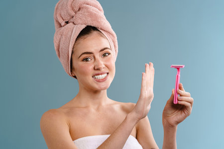 Image of a young displeased woman in towel holding blade and showing no gesture over blue wall backgroundの写真素材