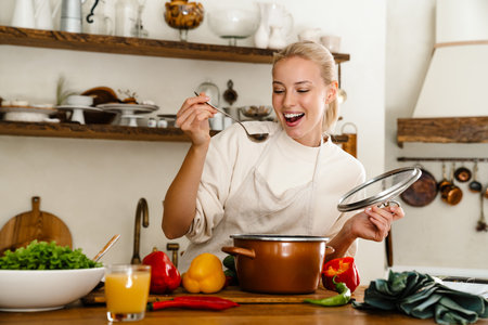 Beautiful pleased woman cooking soup and smiling while making lunch in cozy kitchenの写真素材