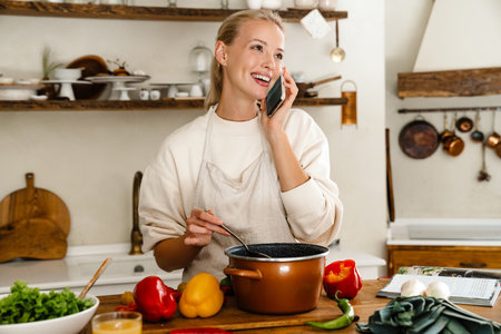 Beautiful happy woman smiling and talking on cellphone while making lunch in cozy kitchenの写真素材