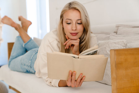 Beautiful focused woman reading book while lying on couch in cozy roomの写真素材