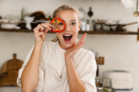 Beautiful delighted woman making fun with vegetables while cooking lunch in cozy kitchenの写真素材