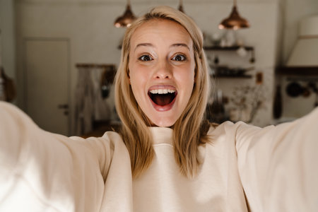 Caucasian excited woman expressing surprise and taking selfie photo while posing at homeの写真素材