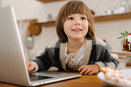 Happy beautiful boy smiling and using laptop while sitting at home kitchenの写真素材