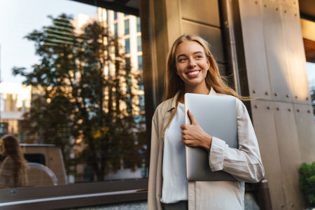 Beautiful young cheerful woman walking outdoors at the city street, carrying laptop computerの写真素材