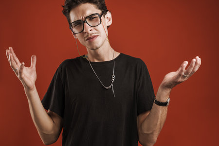 Confused young hipster man standing isolated over red background, shrugging shoulders