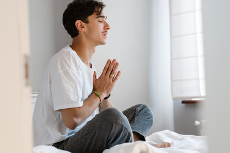 Image of pleased man sitting on bed indoors at home and prayの写真素材
