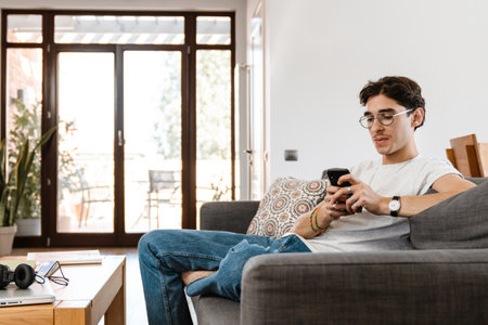 Image of concentrated young handsome man sitting on sofa indoors at home while using mobile phoneの写真素材