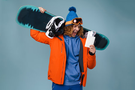 Handsome happy sportsman holding snowboard isolated over blue background, showing flight ticketsの写真素材