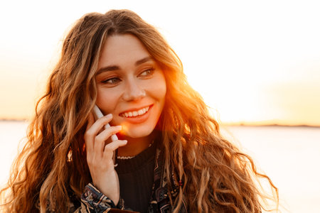 Image of a happy cheery young woman walking outdoors at the beach and talking by smartphone