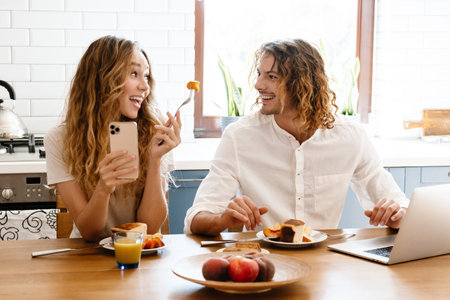 Cheerful beautiful couple using cellphone and laptop while having breakfast in cozy kitchenの写真素材